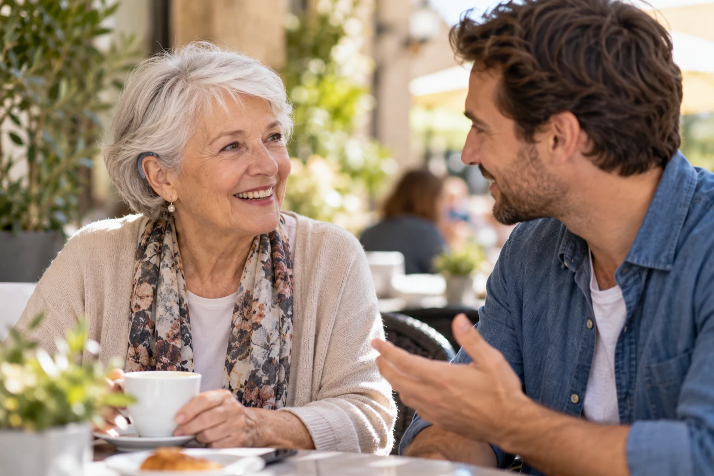 Older woman with small behind-the-ear hearing aid talking with adult grandson outdoors at quiet cafe, candid warm daylight. Illustrative editorial photograph.